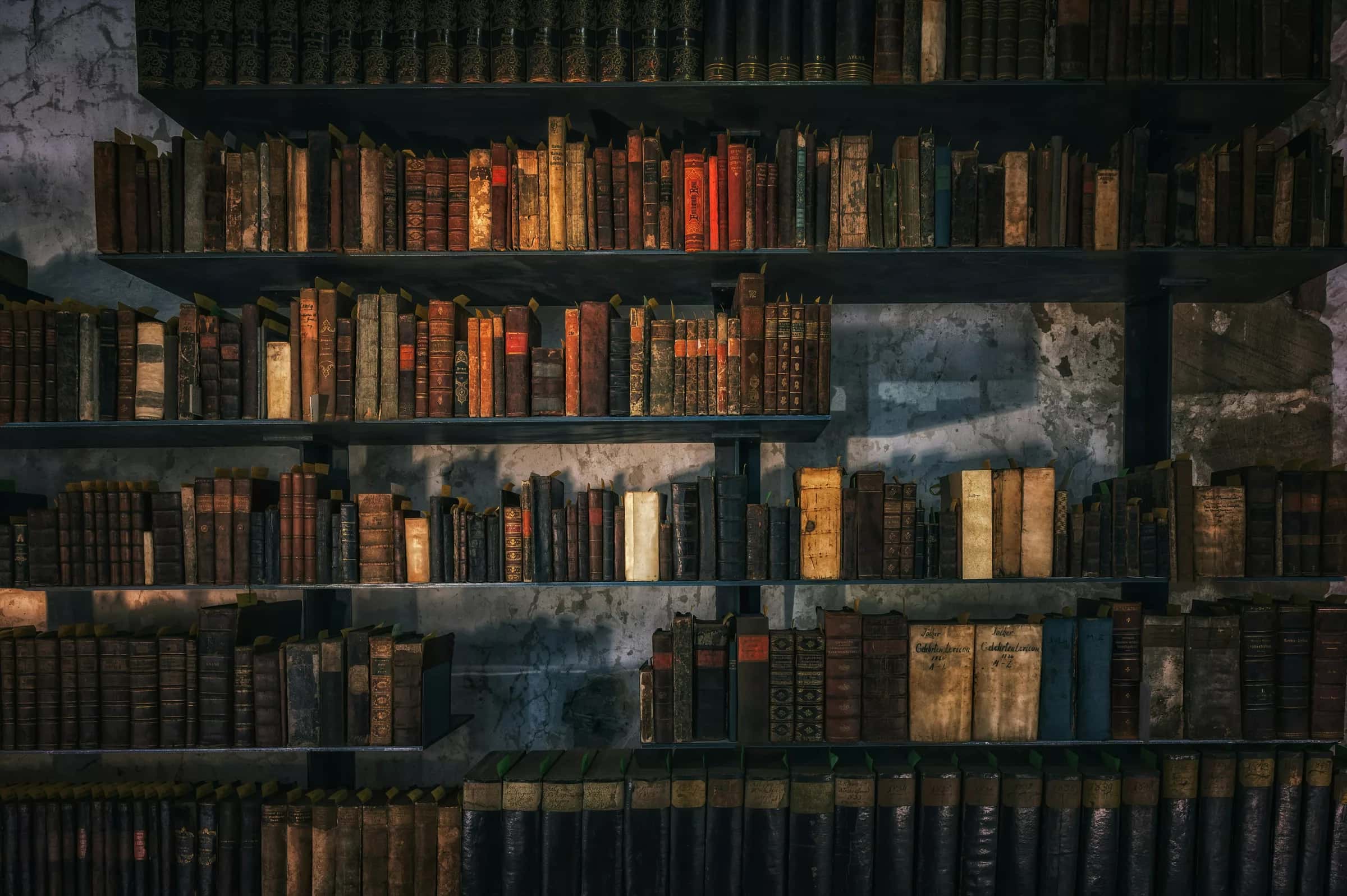 Loş bir kütüphanede raflar dolusu eski, yıpranmış kitap sırtları