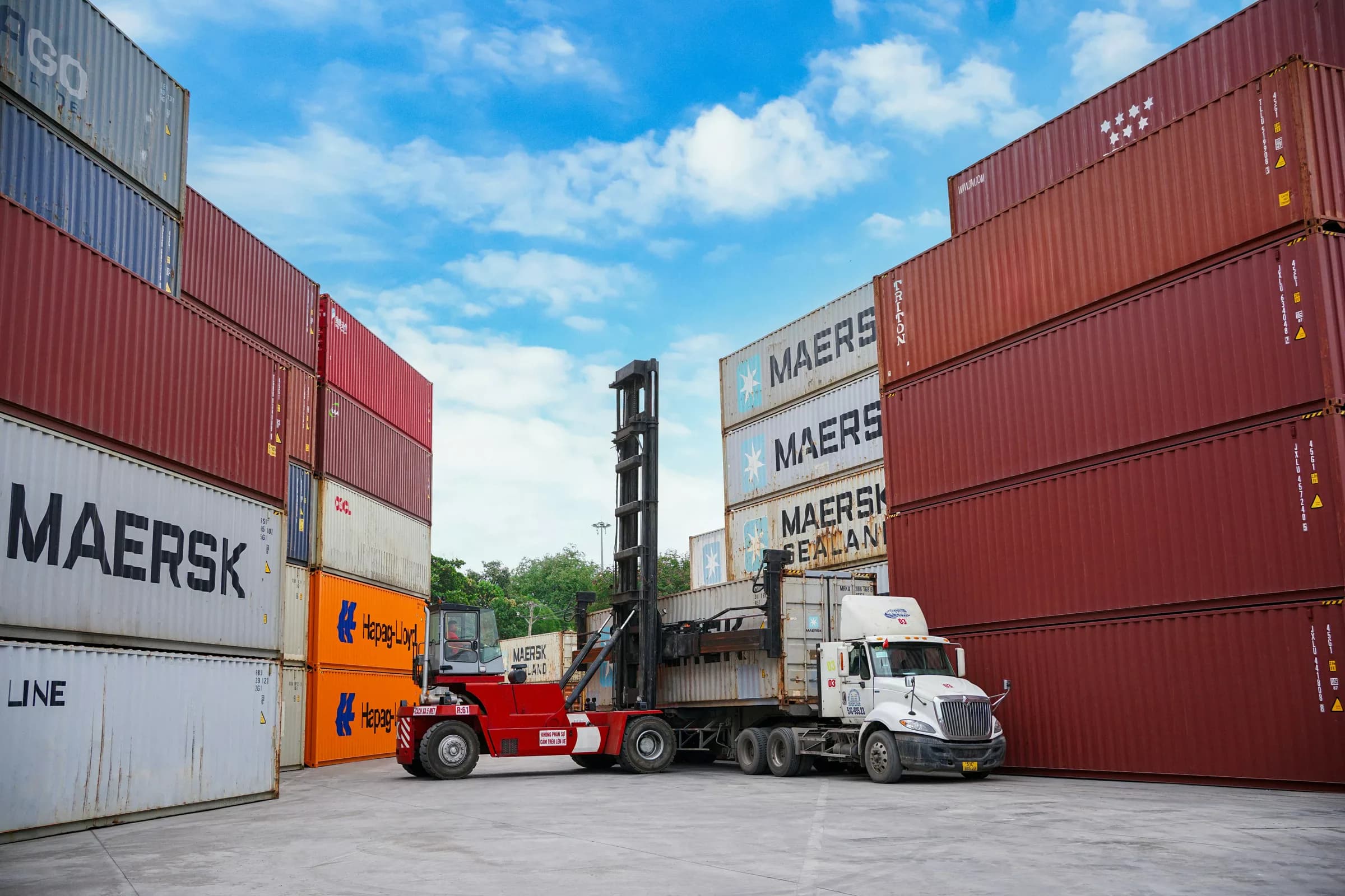 Konteyner sahasında üst üste dizilmiş kutular ve aralarında çalışan bir forklift